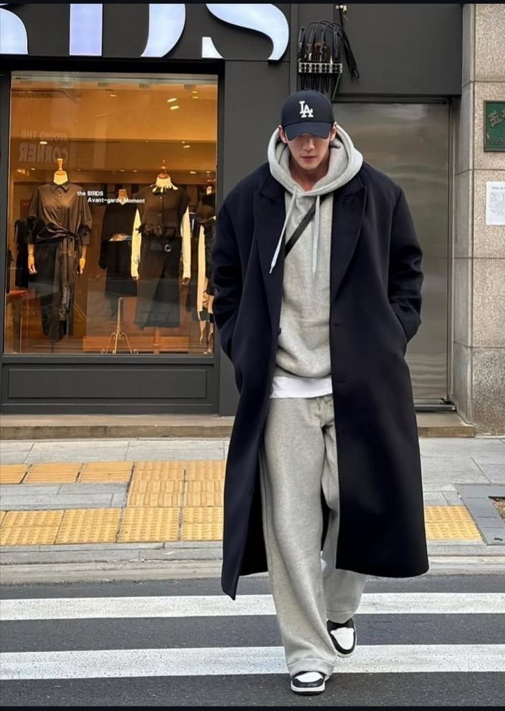 A man walking outdoors wearing a long black coat, grey hoodie, and grey sweatpants.