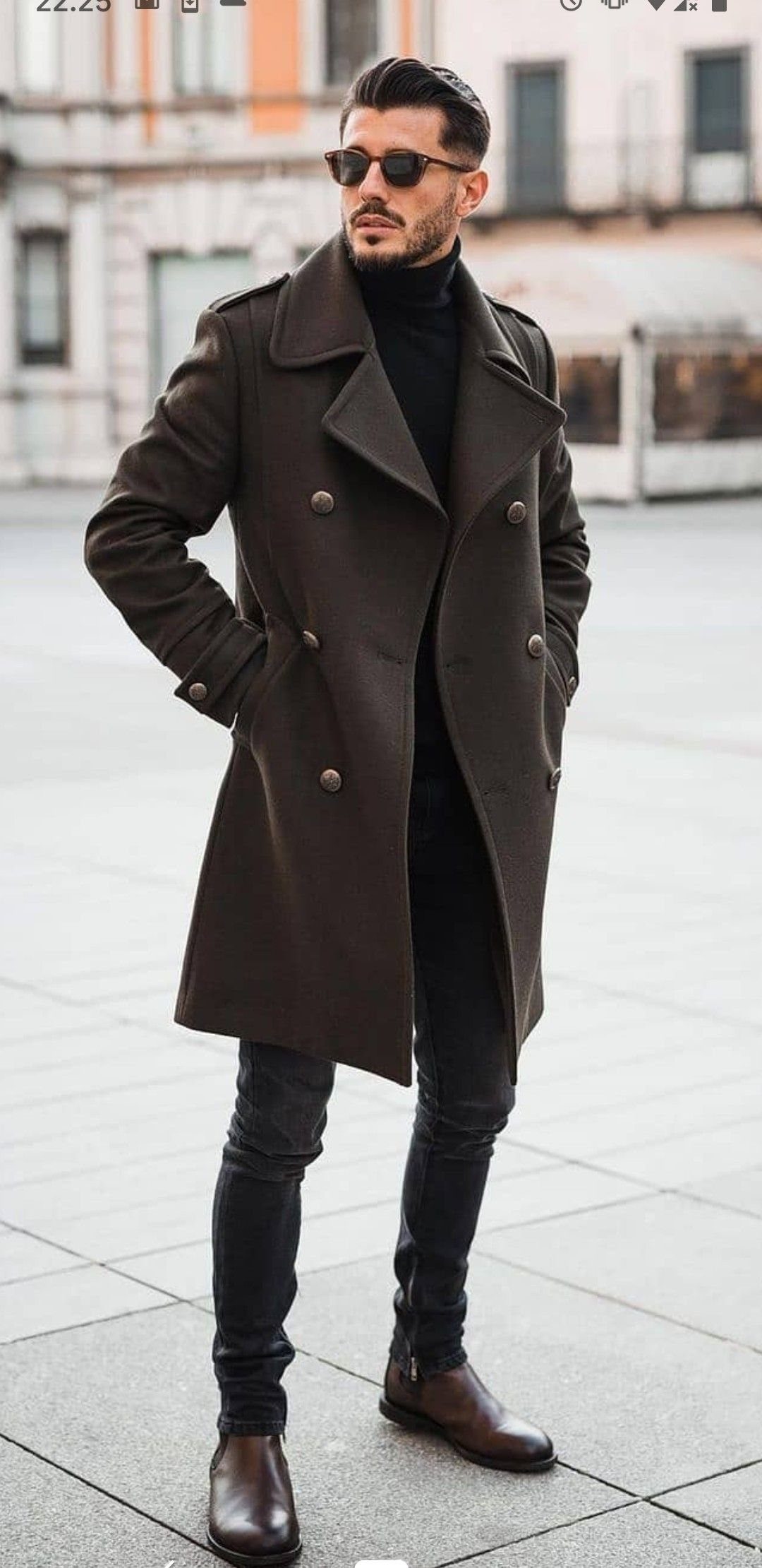 A man posing in a brown wool overcoat, black turtleneck, and dark trousers.