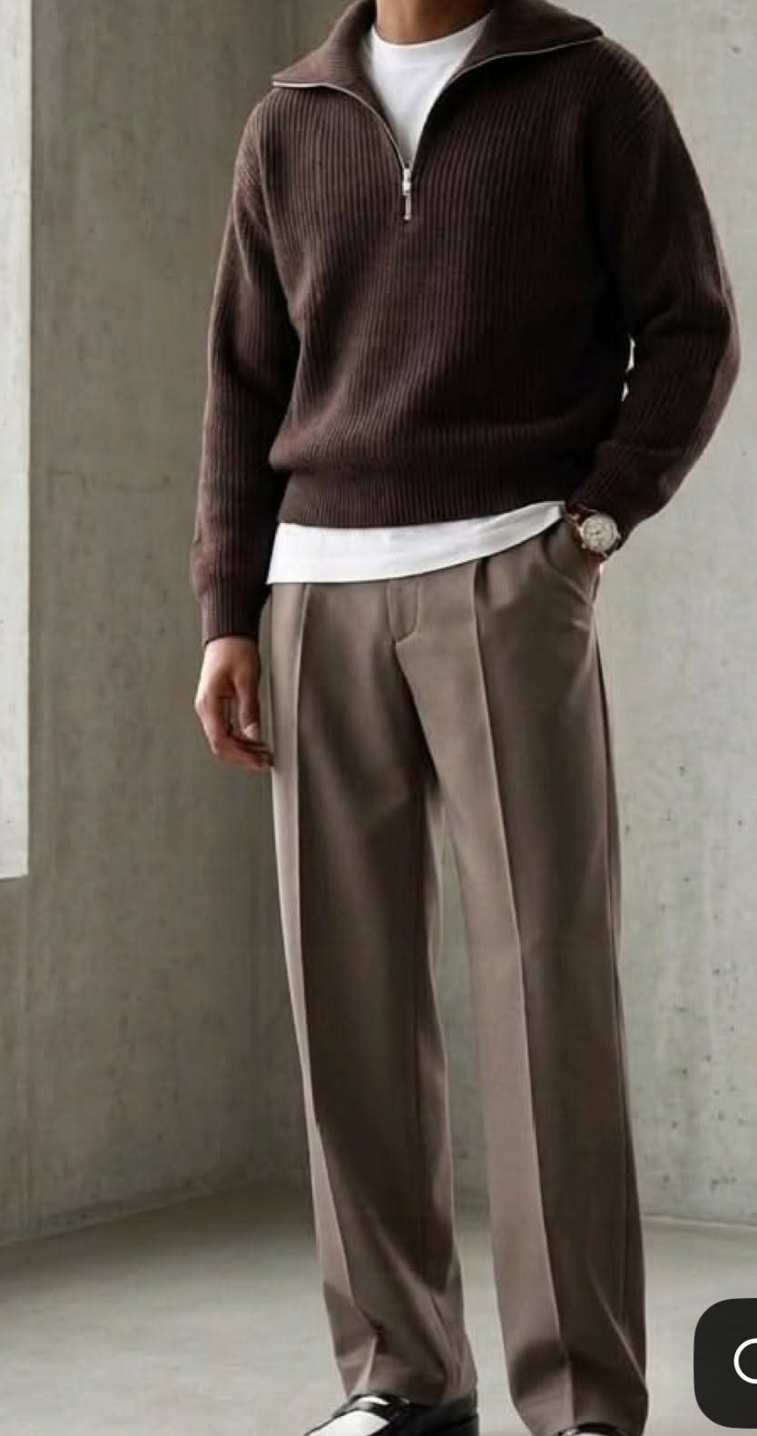 A man wearing a brown ribbed sweater and taupe trousers standing against a grey wall.