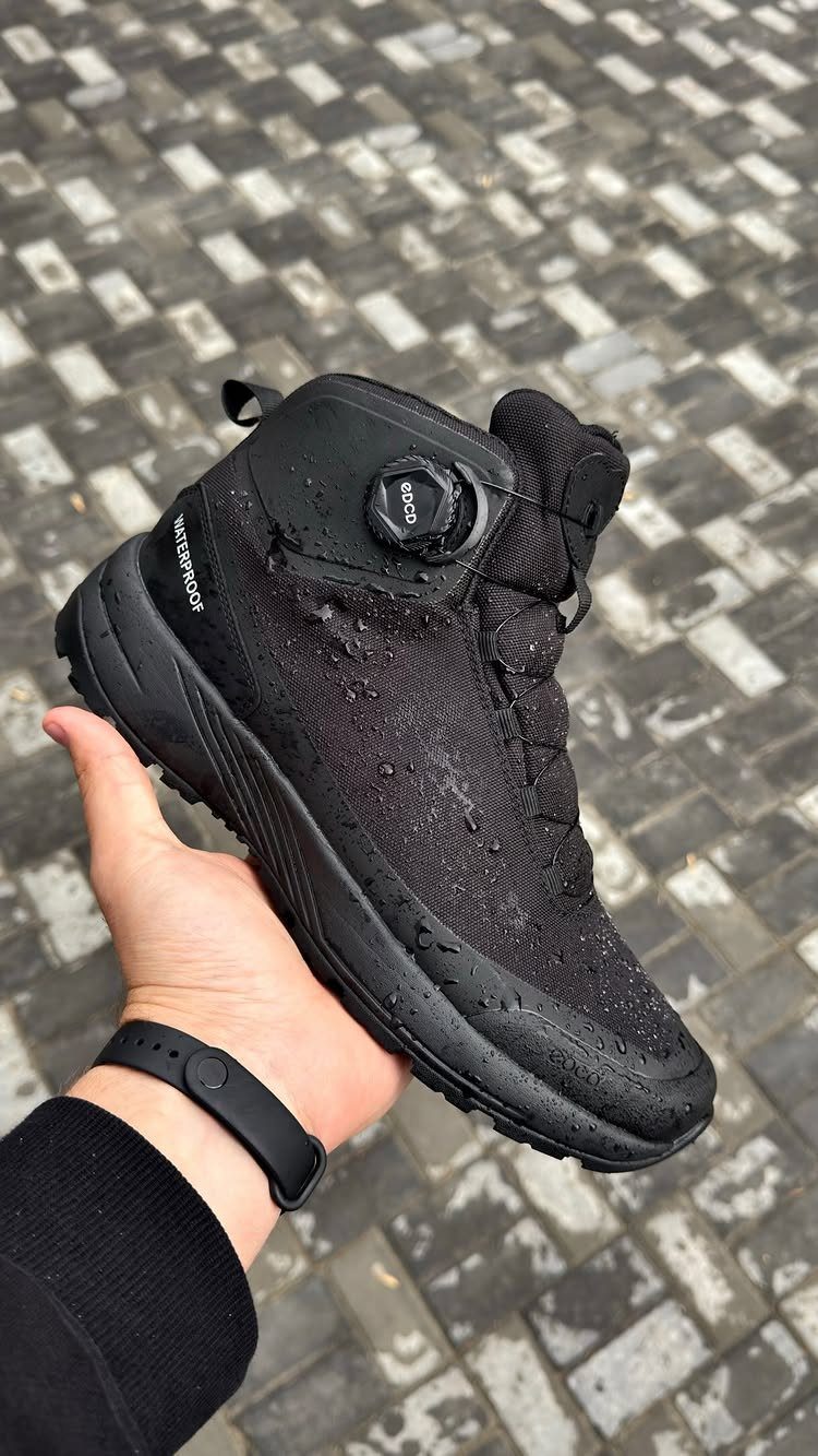 A black waterproof hiking boot with water droplets held by a hand over cobblestones.