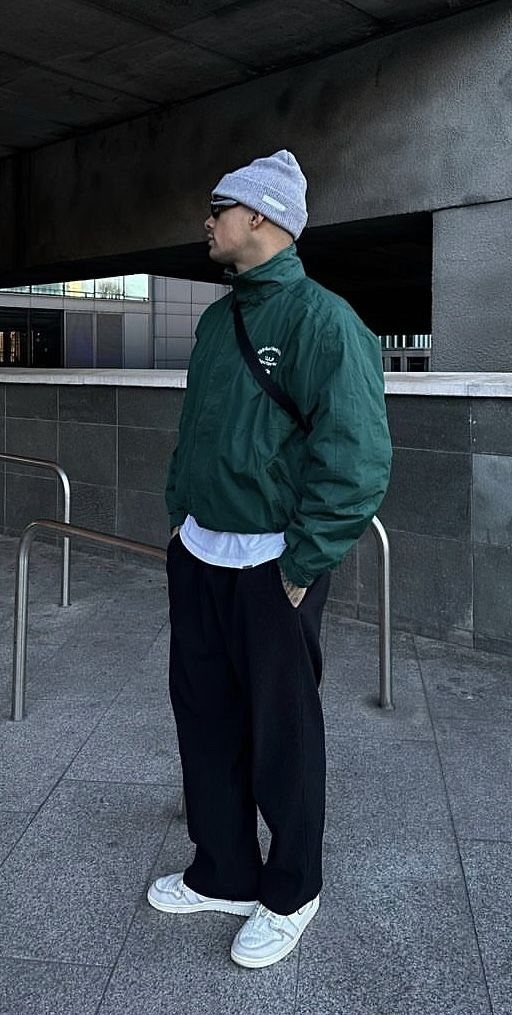 A man wearing a green jacket, black pants, and a grey beanie standing outdoors.